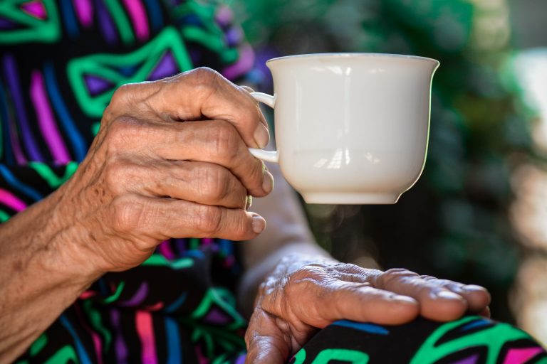 Female elderly hand holding a white cup of coffee and resting other hand on knee wearing a colourful dress