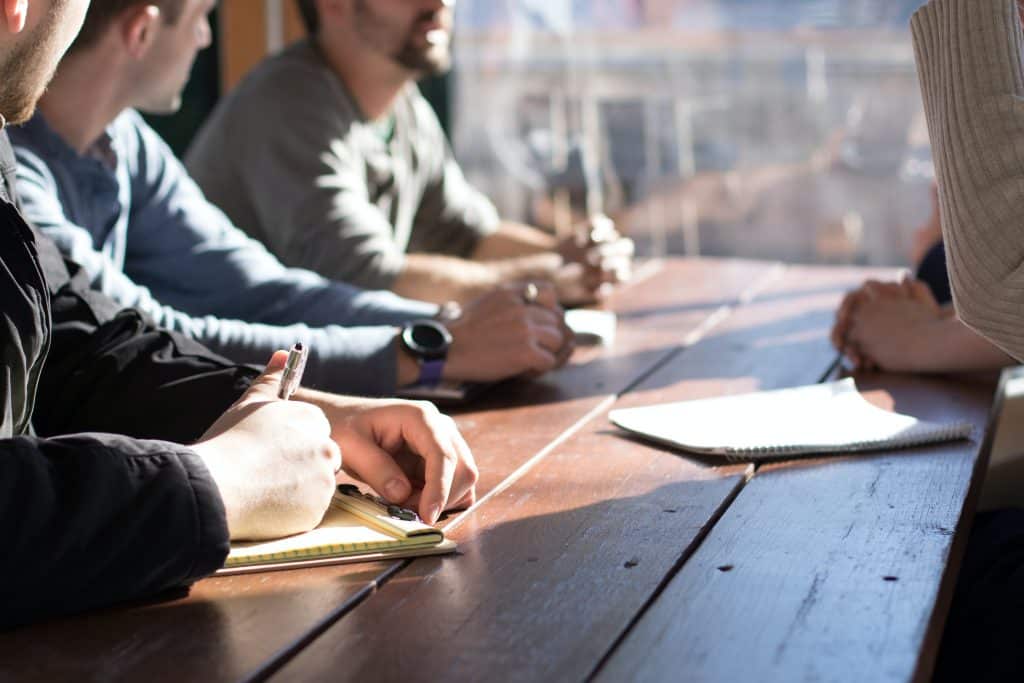 A group of people sit around a wooden table, one has a pen and paper in hand. We only see arms and hands and the wood of the table. Behind the table is a glass window showing a blurred city vista behind.