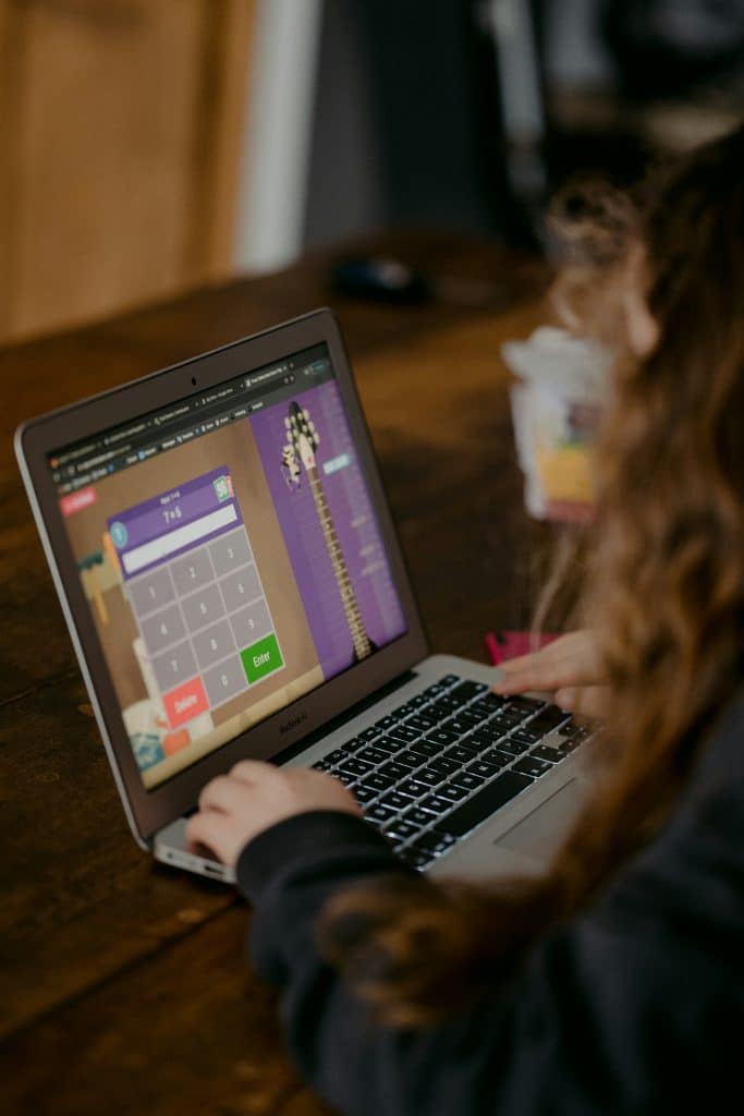 Adult-Only Services And The Children'S Code 10 A grey laptop is open on a wooden desk, showing a calculator on a children's purple and brown background. There is a children's arm, hand and hair visible wearing a black outfit. The photo is taken from the left hand side.