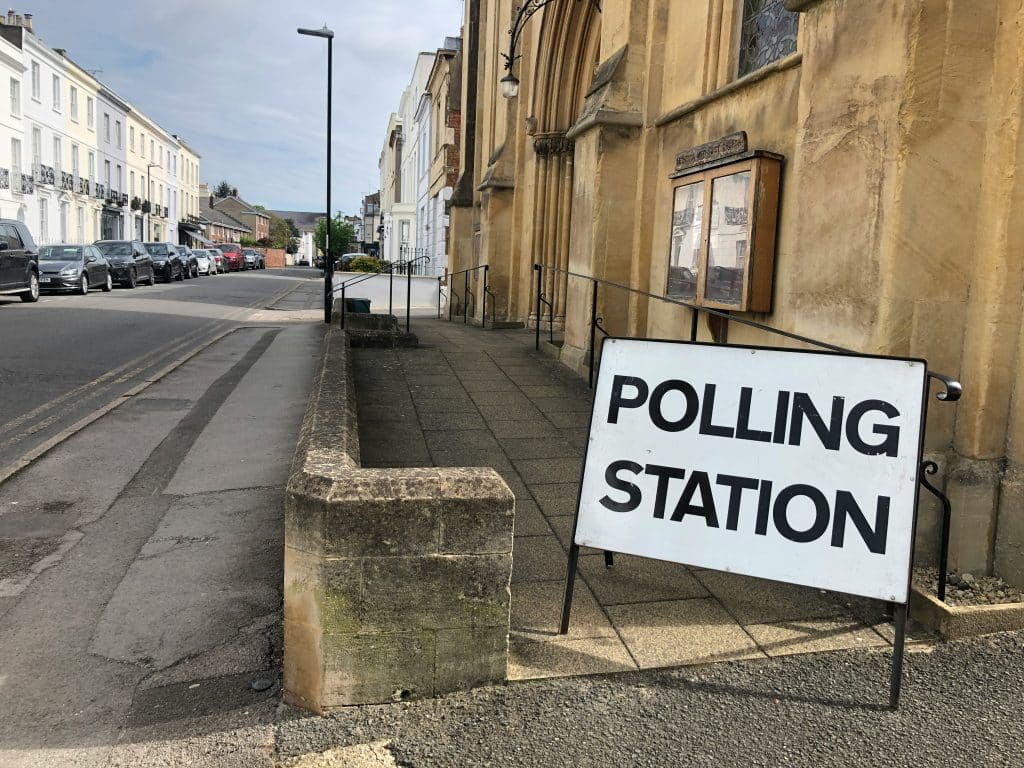 The Election And Data Protection 1 A white sign stands on the corner of a pathway ahead of a large, beige stoned building. It reads 'polling station'. To the left, a row of white terraced houses with cars parked outside can be seen.