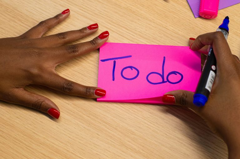 A pair of hands holds a set of pink post it notes and a blue pen, written on the post it notes are the words 'To do'. They are leaning on a desk.