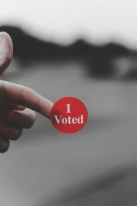 A red sticker with 'I Voted' written in white writing is attached to a finger. The background behind is blurred showing a road.