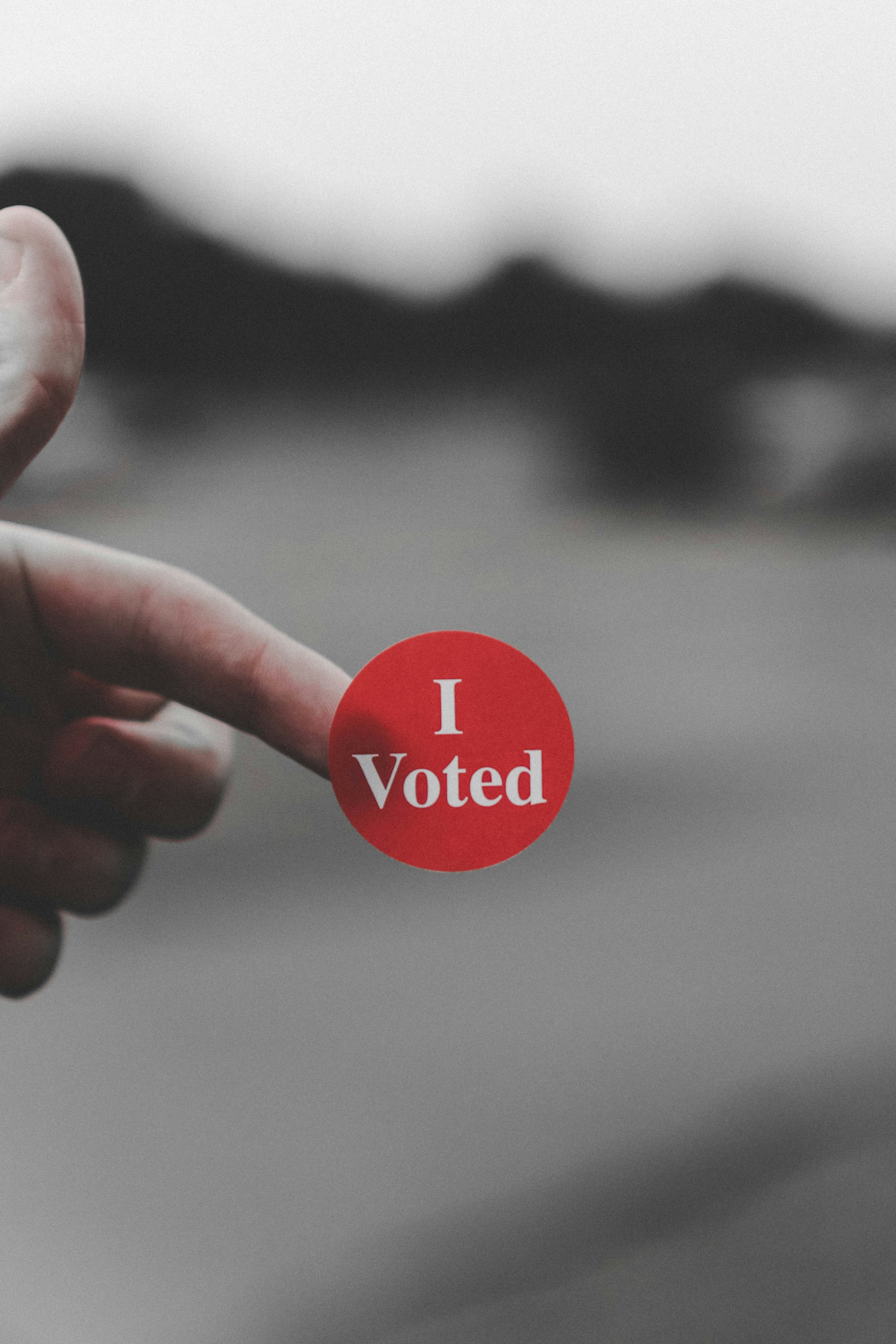 A red sticker with 'I Voted' written in white writing is attached to a finger. The background behind is blurred showing a road.
