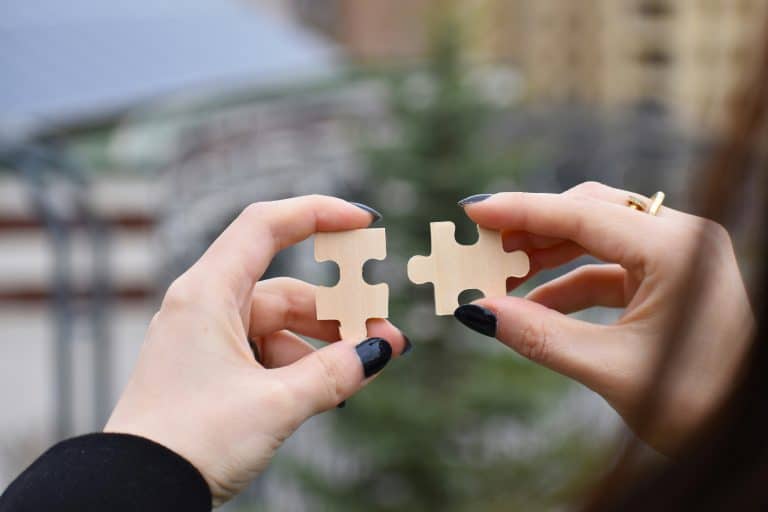 Two hands with black nail varnish hold two pieces of a jigsaw puzzles upright. The pieces of jigsaw are olain and made of wood. A little of the person's hair and cardigan is visible, both are dark coloured.