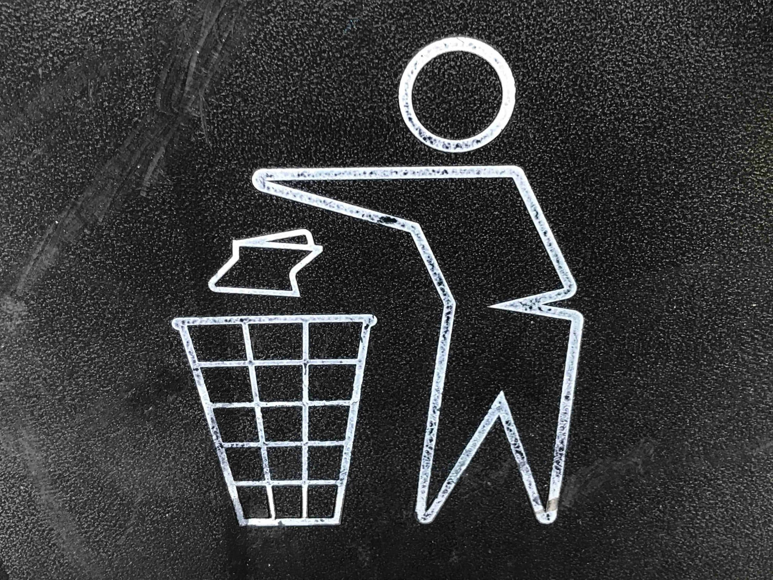 A stick figure is drawn on a dark background using white ink, it appears to show a person throwing a piece of paper into a waste paper bin.