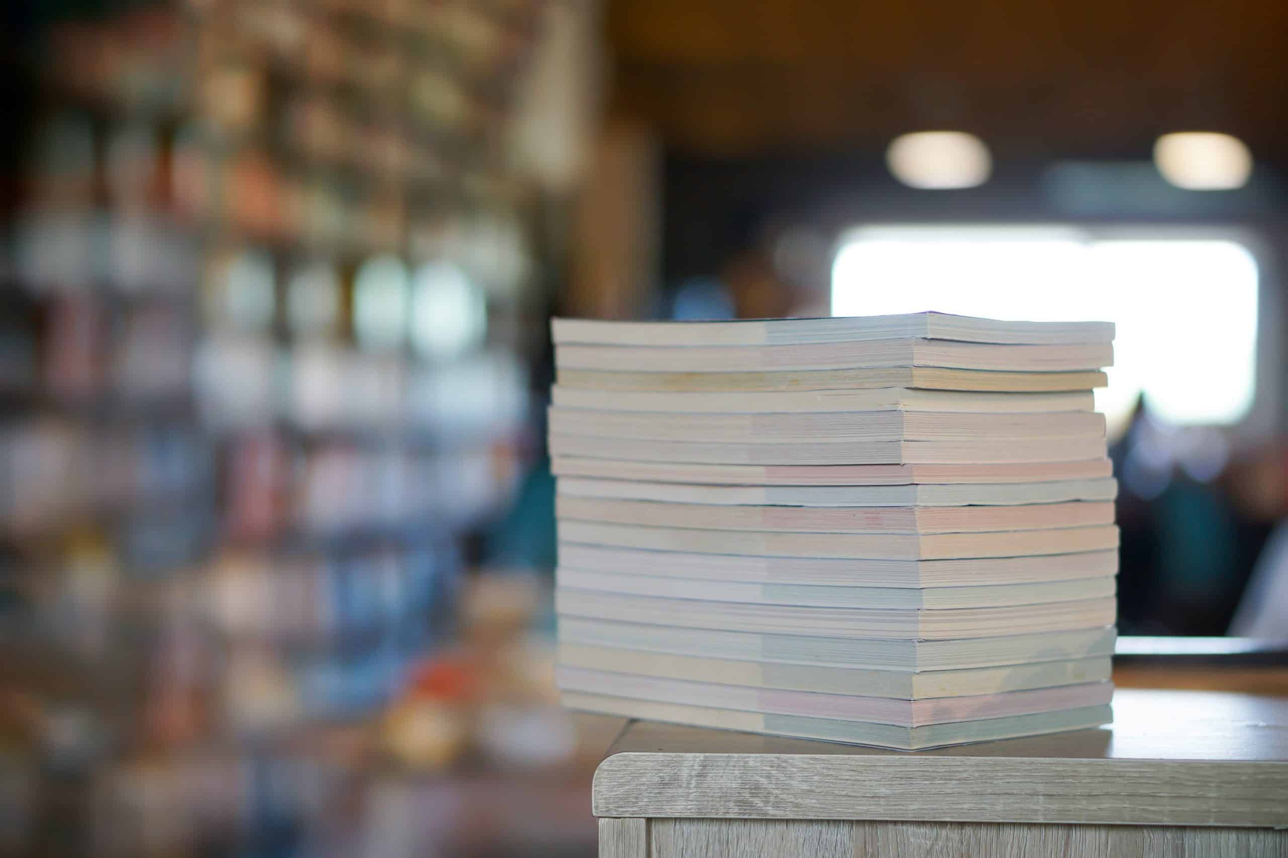 A stack of papers is resting on a table. Out of focus in the background, is a set of floor to ceiling bookcases and a window.