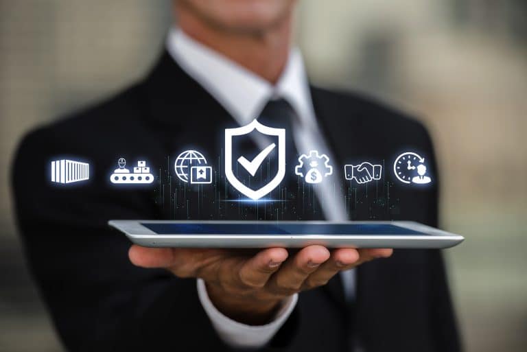 Data protection image from FreePik, image shows a man in a dark coloured suit holding what appears to be an iPad or tablet. Hovering above the tablet are icons depicting security, buildings, people shaking hands and the middle one which is larger than the others, is a shield with a tick in the middle. The icons are in white. The man's hand is in focus but the rest of his body is out of focus.