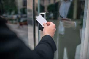 Image shows a man in a black suit using a white ID card on a black scanner box to access a glass door. His image is visible in the reflection. Physical security reviews often consider access and egress of staff, visitors and contractors.