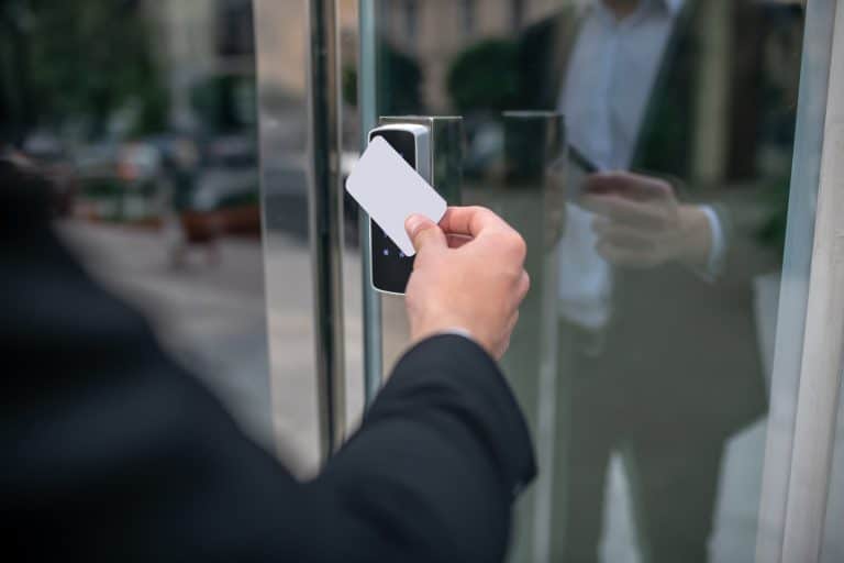 Image shows a man in a black suit using a white ID card on a black scanner box to access a glass door. His image is visible in the reflection. Physical security reviews often consider access and egress of staff, visitors and contractors.