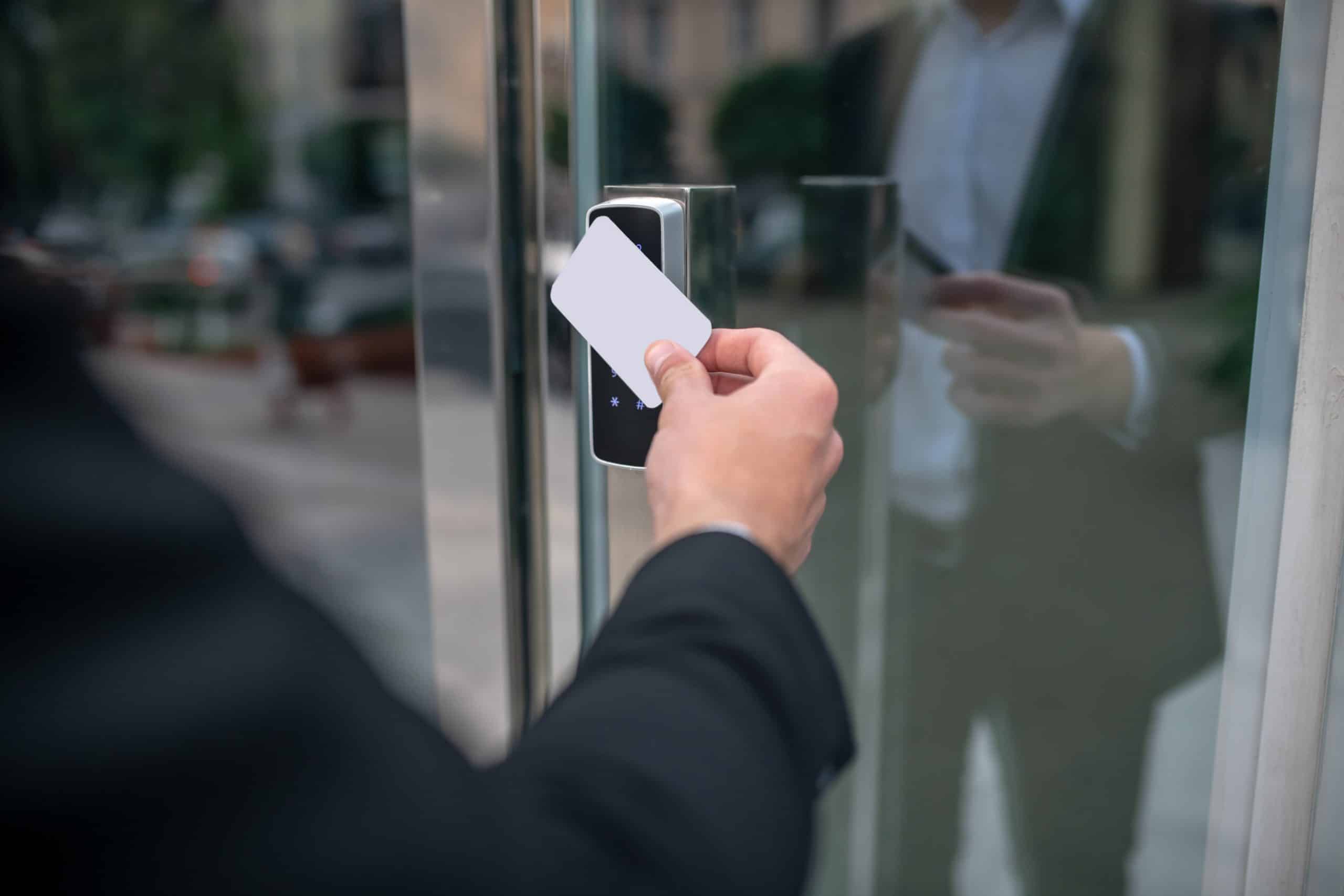Image shows a man in a black suit using a white ID card on a black scanner box to access a glass door. His image is visible in the reflection. Physical security reviews often consider access and egress of staff, visitors and contractors.