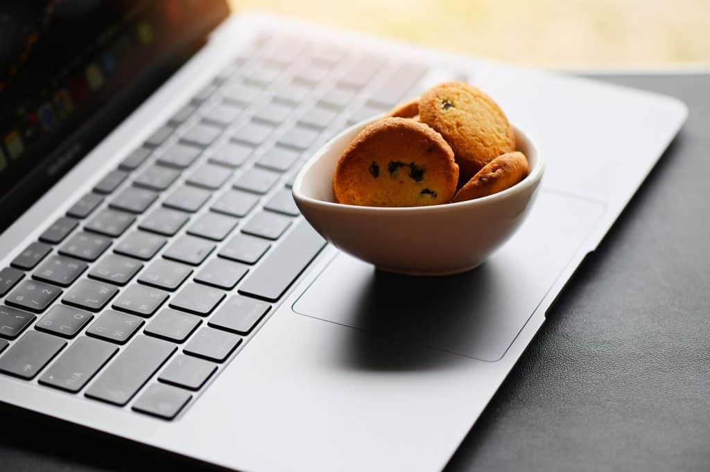 The image shows a grey laptop keyboard resting on a grey table, the screen of the laptop is only slightly visible in the corner of the image. On top of the keyboard, where the mousepad is, is a white bowl with a handful of small chocolate chip cookies in, demonstrating cookies and data protection.