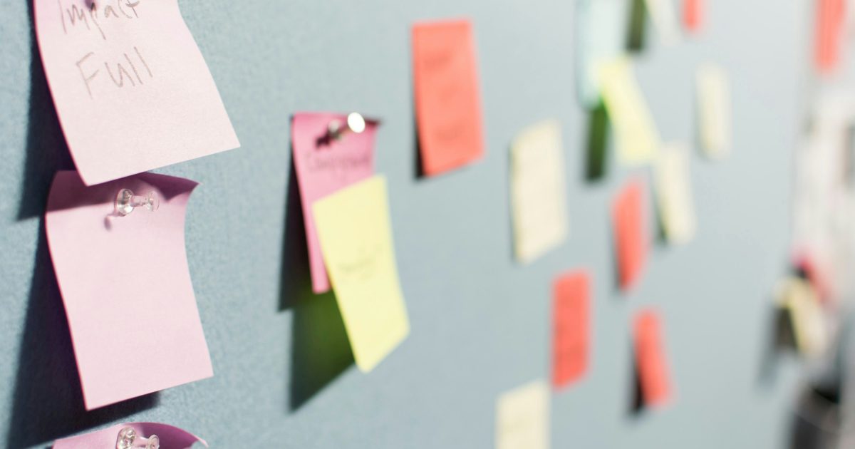Image shows colourful post-it notes fixed to a grey pinboard with drawing pins. They are mostly out of focus. The nearest ones to the camera are pink and one of them has 'Impactful' written on it in black ink.